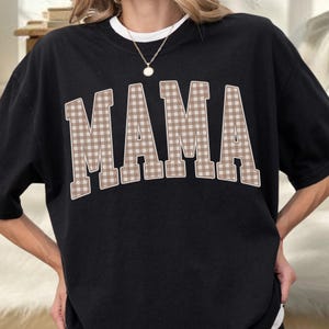 May include: A black short-sleeved t-shirt featuring the word MAMA printed in large, block-style letters across the chest. The letters have a light brown and white gingham check pattern with a thin white outline, and a gold-toned necklace is worn.