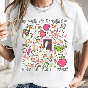 May include: A white t-shirt featuring a colorful graphic print of multiple cartoon ducks engaged in school-related activities. The text "most definitely" appears at the top and "not all in a row" at the bottom, surrounding illustrations of books, pencils, an apple, and an A+ star.