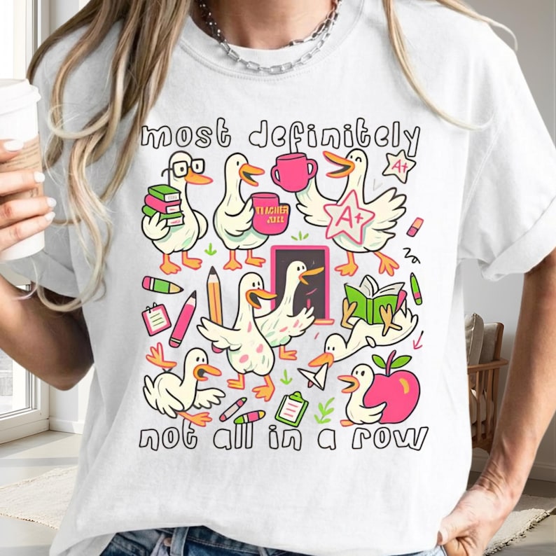 May include: A white t-shirt featuring a colorful graphic print of multiple cartoon ducks engaged in school-related activities. The text "most definitely" appears at the top and "not all in a row" at the bottom, surrounding illustrations of books, pencils, an apple, and an A+ star.
