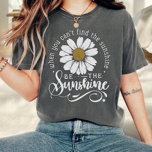 A person wearing a gray t-shirt with a white flower design and the text "When you can't find the sunshine, be the sunshine" printed on it.