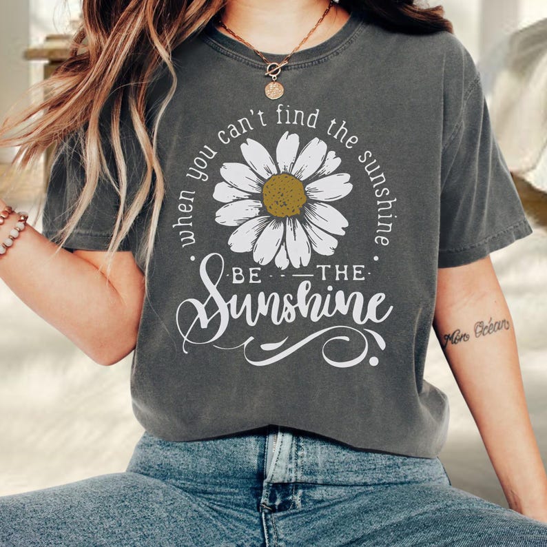 A person wearing a gray t-shirt with a white flower design and the text "When you can't find the sunshine, be the sunshine" printed on it.