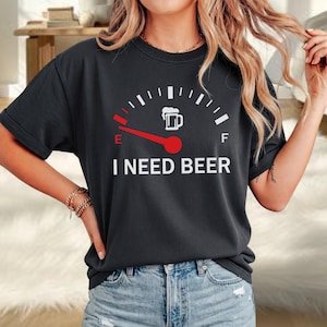 Könnte beinhalten: Ein dunkelgraues Kurzarm-T-Shirt mit einer weiß-roten Grafik einer Tankanzeige. Die Nadel zeigt auf E, und ein weißes Bierkrug-Symbol befindet sich in der Nähe von F. Der Text I NEED BEER ist in weißen Buchstaben unter der Anzeige aufgedruckt.