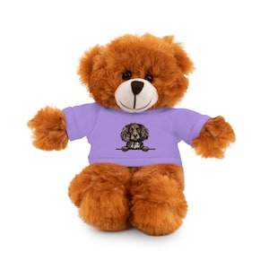 May include: A brown teddy bear wearing a purple t-shirt with a cartoon image of a brown and white dog peeking out from behind the shirt.