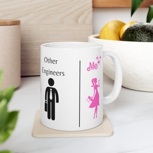 May include: White ceramic mug with the text "Other Engineers" and a stick figure in a suit and tie. The other side of the mug has the word "Me" and a pink stick figure of a woman. The mug is on a beige coaster.