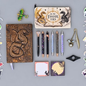 May include: A collection of stationery items featuring dragon motifs, including a textured brown journal, a zippered pencil case with the text In My Fantasy Era, five pens, a metal feather bookmark, two sticky note pads, and various dragon stickers.