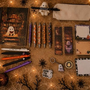May include: A collection of Halloween-themed stationery including a spiral notebook, five patterned pens, a white pencil case, two bookmarks, three sticky note pads, a ghost figurine, a keychain, and several ghost-themed stickers on a textured surface.