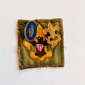 May include: A vintage Boy Scout patch with gold and brown embroidered details, featuring a fleur-de-lis emblem and the words "BE PREPARED". A blue and gold pin with a hand gesture is also present.