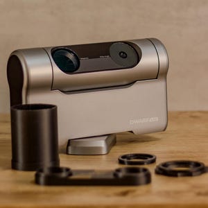 May include: A silver 3D camera with a black lens and the word "DWARF3D" printed on the side. The camera is sitting on a wooden table with a black lens cap and three black rings.