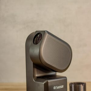 May include: A grey projector with a lens on the front and a black base. The projector is sitting on a wooden table with several small black metal parts scattered around it.