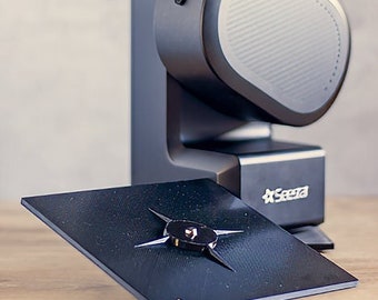 May include: A black and gray 3D printer with a small black and gold plate on the front. The plate has the text "VO" and a number "1" on it. The printer is on a wooden surface.