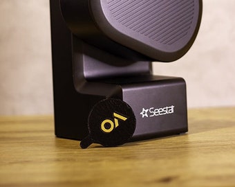 May include: A black projector with a round lens and a textured speaker grill. The projector has a black base with the text "Seestar" printed on it.