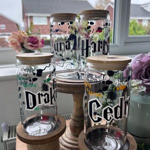 May include: Four glass jars with wooden lids and personalised Harry Potter themed designs. The jars are on wooden stands and feature the names Luna, Harry, Drake, and Cedric.