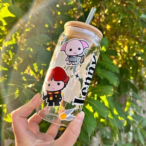May include: A clear glass water bottle with a bamboo lid and a straw. The bottle has several Harry Potter themed stickers, including Harry Potter, Hermione Granger, and Dobby. The bottle is labelled "Lenny".