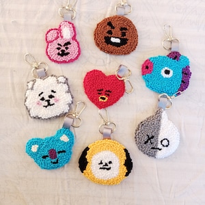 May include: Eight colorful embroidered keychains featuring characters from the popular South Korean boy band, BTS. The keychains are made of yarn and feature different characters, including a brown bear, a white dog, a blue bird, a pink bunny, a red heart, a gray and white panda, a yellow dog, and a blue koala.