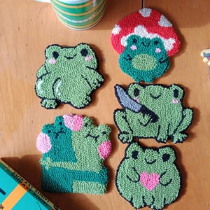 May include: Five green frog-shaped coasters with different designs. One frog is under a red mushroom, one frog is holding a knife, one frog is holding a pink heart, and two frogs are hugging each other. The coasters are made of yarn and are a fun and whimsical addition to any home.