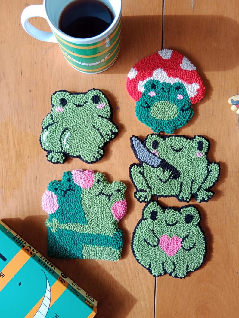 May include: Five green frog-shaped coasters with different designs. One frog is under a red mushroom, one frog is holding a knife, one frog is holding a pink heart, and two frogs are hugging each other. The coasters are made of yarn and are a fun and whimsical addition to any home.