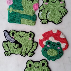 May include: Five green frog-shaped felt ornaments with different expressions. One frog is holding a knife, another is holding a heart, and another is wearing a red mushroom hat.