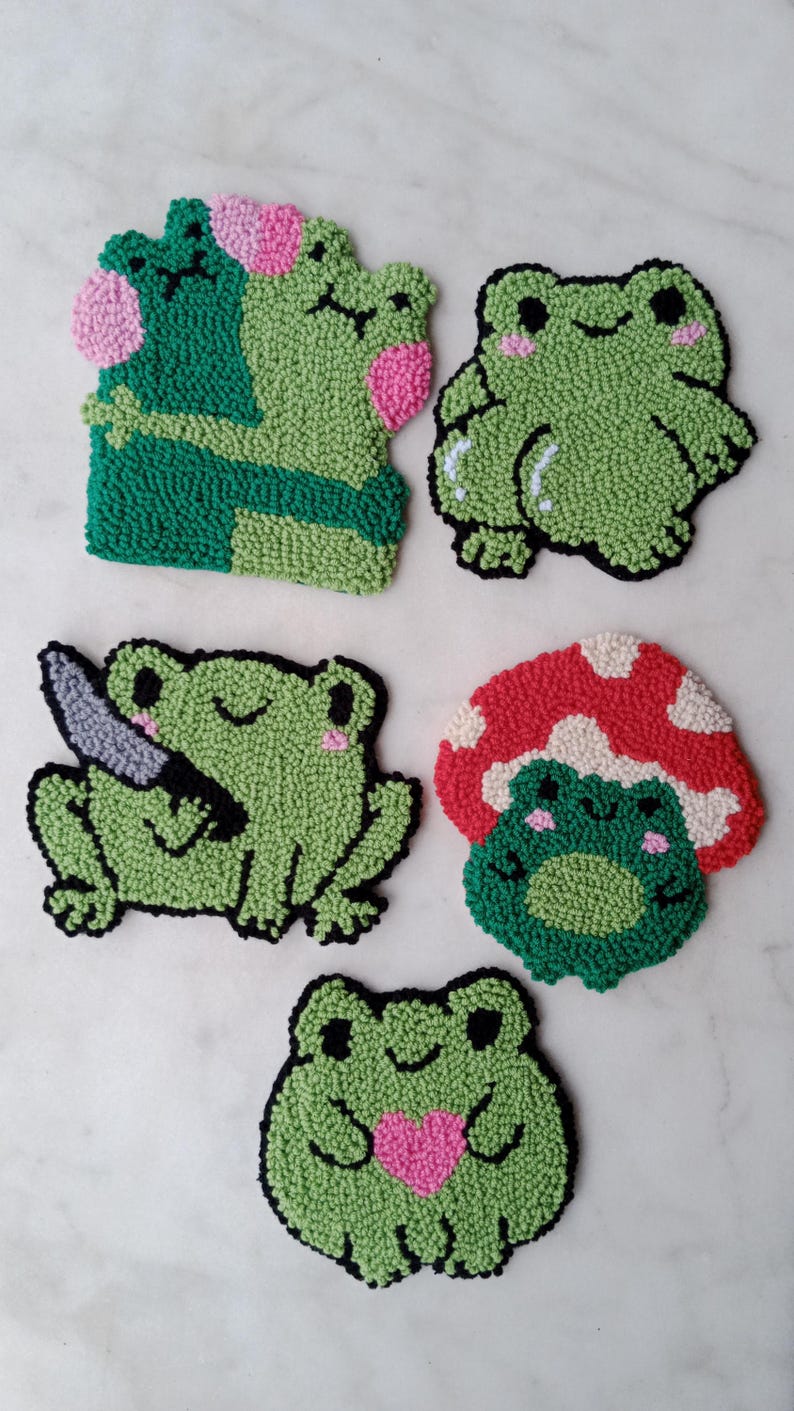 May include: Five green frog-shaped felt ornaments with different expressions. One frog is holding a knife, another is holding a heart, and another is wearing a red mushroom hat.