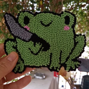 May include: A green frog with black eyes and a pink blush holding a grey knife. The frog is made of yarn and is a needle felted craft.