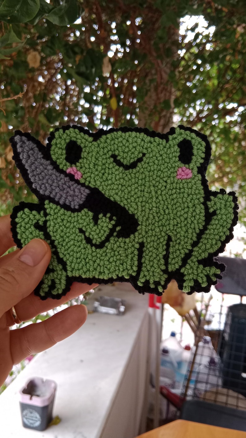 May include: A green frog with black eyes and a pink blush holding a grey knife. The frog is made of yarn and is a needle felted craft.