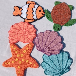 May include: A collection of colorful, handmade, sea-themed coasters. The set includes a fish, a turtle, a starfish, and three different seashell designs. The coasters are made with a textured, looped yarn technique, and are in shades of orange, green, brown, pink, purple, and blue.