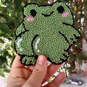 May include: A green felt frog with black eyes and pink cheeks. The frog is stitched with a fluffy texture.