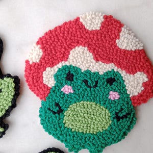 May include: A green frog with a red and white mushroom hat. The frog has black eyes and a pink blush on its cheeks. The frog is made of yarn and is a handmade craft.