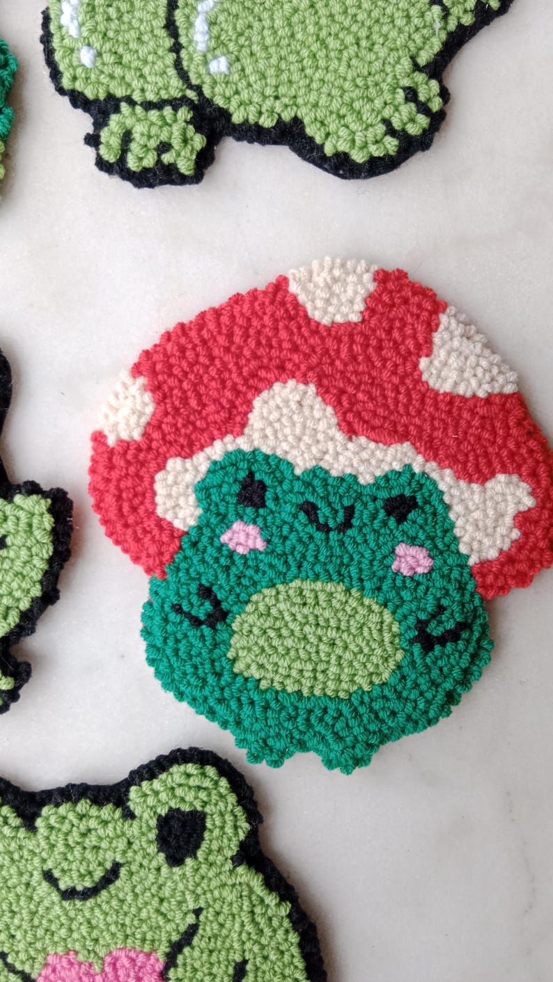 May include: A green frog with a red and white mushroom hat. The frog has black eyes and a pink blush on its cheeks. The frog is made of yarn and is a handmade craft.