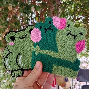 May include: Three green felt frog plushies with pink cheeks. The frogs are hugging each other.
