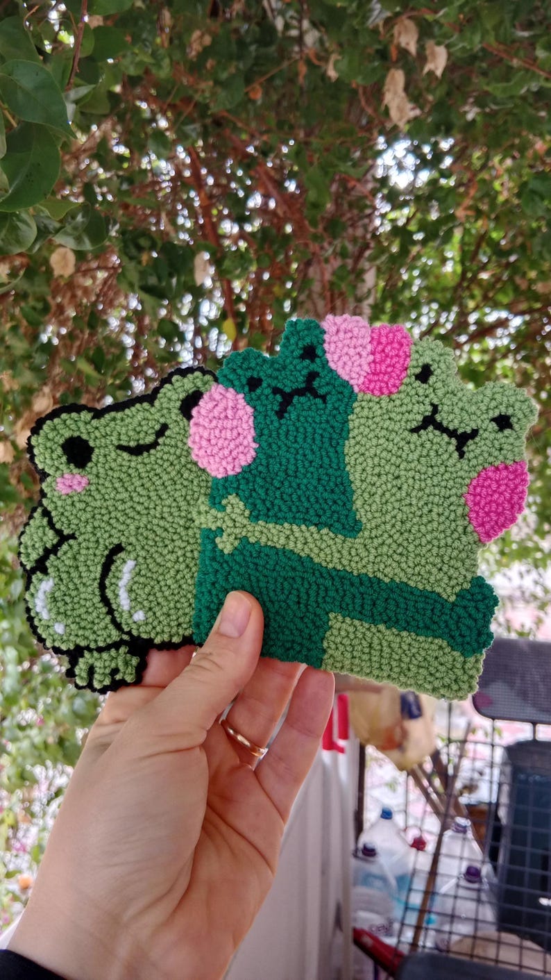 May include: Three green felt frog plushies with pink cheeks. The frogs are hugging each other.