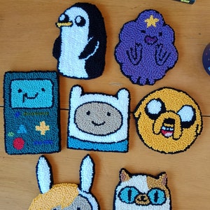 May include: A set of seven colorful felt character patches featuring Finn the Human, Jake the Dog, Lumpy Space Princess, BMO, Marceline the Vampire Queen, Bubblegum, and Ice King from the animated television series Adventure Time.
