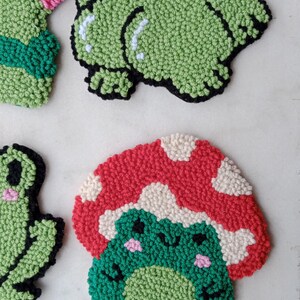 May include: Four handmade felt frog ornaments. One frog is green with black eyes and pink cheeks. Another frog is green with black eyes and pink cheeks and is sitting on a red and white mushroom. The other two frogs are green with black eyes and pink cheeks.