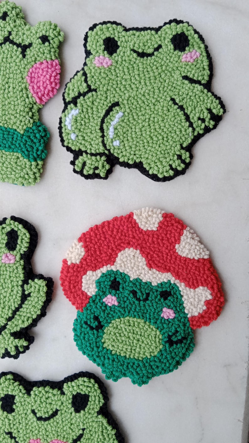 May include: Four handmade felt frog ornaments. One frog is green with black eyes and pink cheeks. Another frog is green with black eyes and pink cheeks and is sitting on a red and white mushroom. The other two frogs are green with black eyes and pink cheeks.