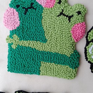 May include: Two green felt frog plushies with pink cheeks and black eyes. One frog is hugging the other frog.