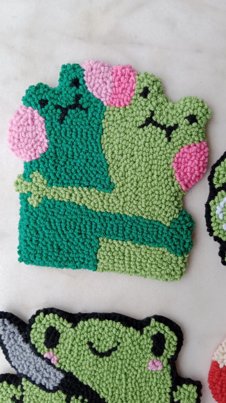 May include: Two green felt frog plushies with pink cheeks and black eyes. One frog is hugging the other frog.