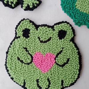 May include: Green felt frog holding a pink heart. The frog has black eyes and a black outline.