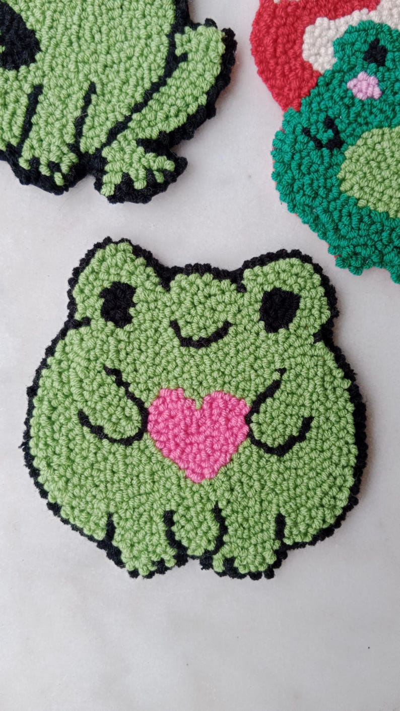 May include: Green felt frog holding a pink heart. The frog has black eyes and a black outline.