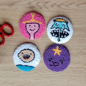 May include: Four round, colorful coasters featuring cartoon characters. The coasters are made of a textured material and have a white, pink, blue, and purple color scheme. Each coaster has a different character design.