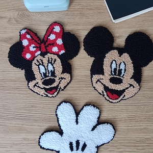 May include: Three embroidered patches featuring cartoon characters. One patch depicts Minnie Mouse with a red polka dot bow, another Mickey Mouse, and a white glove with black details. The patches are on a wooden surface.