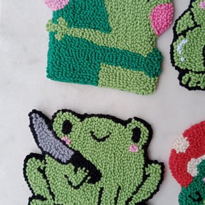 May include: A set of five handmade felt frog ornaments. The frogs are green with black outlines and pink accents. One frog is holding a knife, another is hugging another frog, and the other three are smiling.