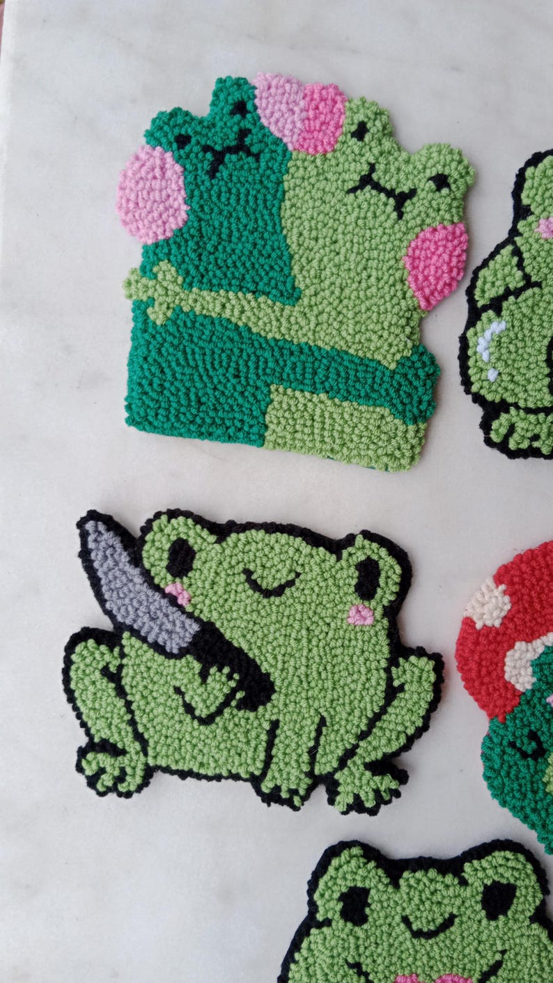May include: A set of five handmade felt frog ornaments. The frogs are green with black outlines and pink accents. One frog is holding a knife, another is hugging another frog, and the other three are smiling.