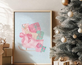 May include: A framed watercolor print featuring a pink sleigh filled with wrapped gifts in pink, green, and beige. The sleigh has a large pink bow and sits on a light blue background with falling snowflakes. A Christmas tree with ornaments is in the background.