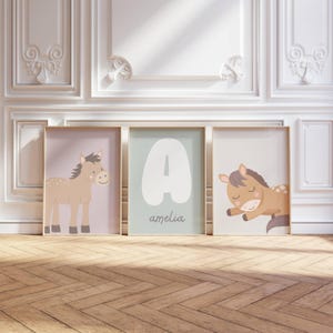 May include: Three framed art prints with a horse theme. One print shows a standing horse, another displays the letter "A" with the name "amelia", and the third depicts a sleeping horse. The prints have gold frames and are set against a white wall.