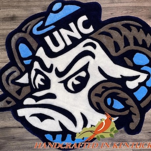 May include: A blue and white rug with a cartoon ram wearing a blue hat with the letters UNC on it. The rug is handcrafted in Kentucky.