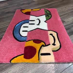 Puede incluir: Una alfombra rosa con un dise&ntilde;o de dibujos animados que presenta un hongo con acentos rojos y amarillos, un hongo azul y blanco y un hongo verde y blanco.