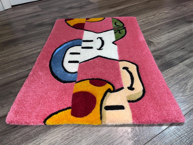 Puede incluir: Una alfombra rosa con un dise&ntilde;o de dibujos animados que presenta un hongo con acentos rojos y amarillos, un hongo azul y blanco y un hongo verde y blanco.