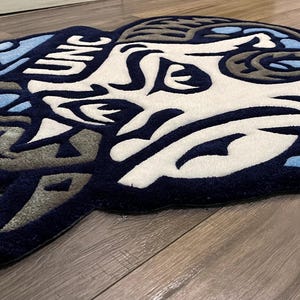 May include: A blue and white rug with a stylized ram's head design. The rug has the letters "UNC" in the upper left corner.