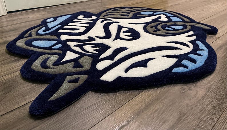 May include: A blue and white rug with a stylized ram's head design. The rug has the letters "UNC" in the upper left corner.