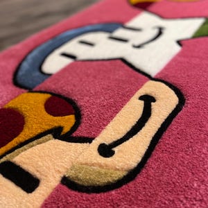 Puede incluir: Una alfombra rosa con un dise&ntilde;o de personaje de dibujos animados. El personaje es una figura blanca con un sombrero azul y un hongo amarillo. El personaje sonr&iacute;e y tiene un contorno negro.