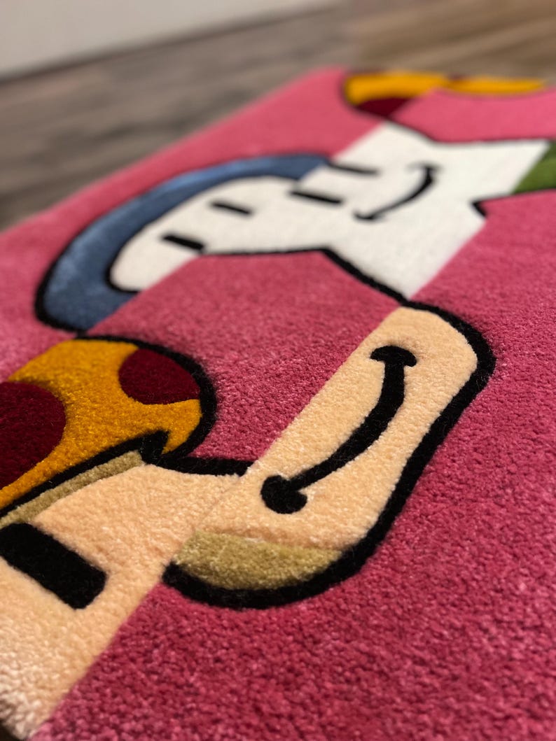 Puede incluir: Una alfombra rosa con un dise&ntilde;o de personaje de dibujos animados. El personaje es una figura blanca con un sombrero azul y un hongo amarillo. El personaje sonr&iacute;e y tiene un contorno negro.